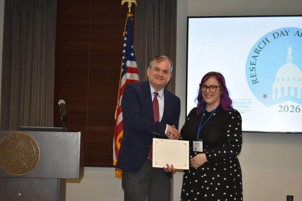 A man and woman shake hands as she receives a certificate during a Research Day award presentation, with an Oklahoma state seal podium and U.S. flag nearby.