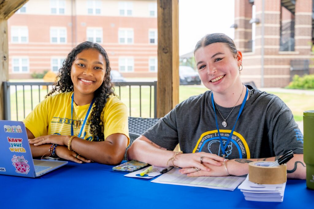 Two students wearing event lanyards sit behind a table outdoors, smiling toward the camera while assisting with a campus welcome or check-in activity.