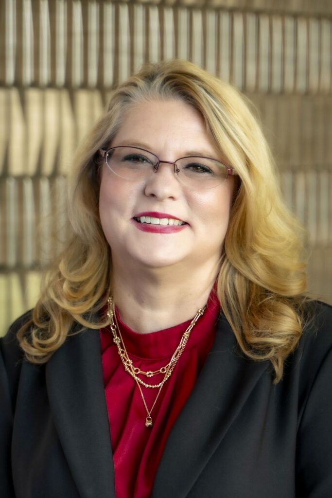 Christina Smith smiles for a professional headshot, wearing glasses, a red blouse, black blazer, and layered gold necklaces.