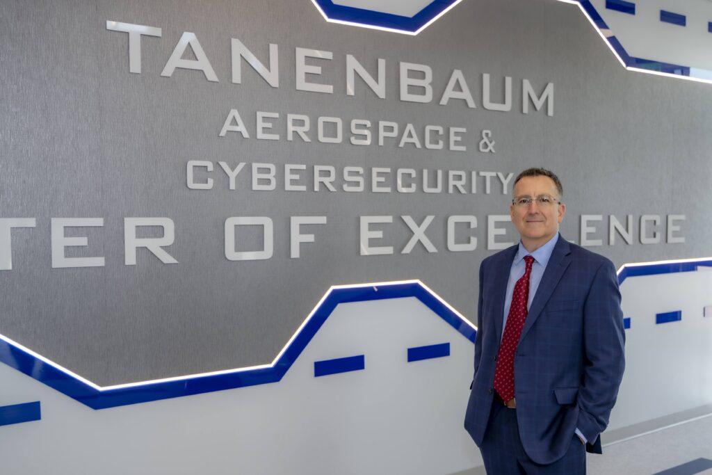 Man in a blue suit and red tie standing beside a wall sign for the Tanenbaum Aerospace & Cybersecurity Center of Excellence.