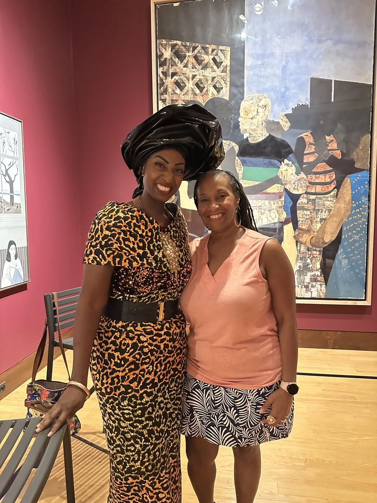 Two women smiling together in an art gallery, standing in front of a large mixed-media artwork.
