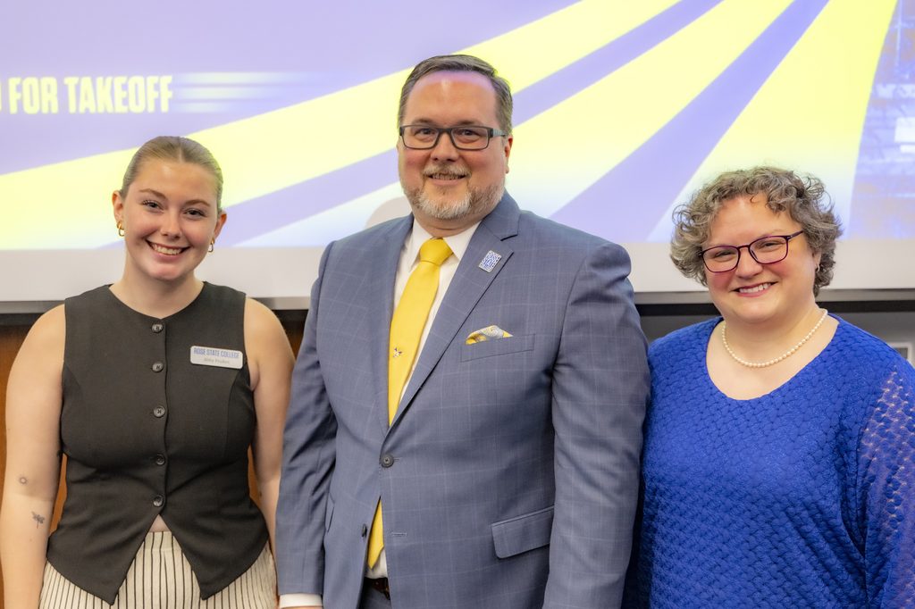 Three people smiling together at Rose State College’s State of Rose State event, with a man in a blue suit and yellow tie standing between two women in front of a Cleared for Takeoff presentation screen.
